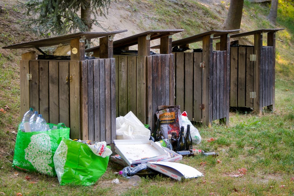 A row of four outdoor wooden waste bins with slanted roofs, positioned on a grassy area near a woodland setting, with some of the bins showing signs of weathering and slight discoloration. In front of the bins, there are various discarded items including plastic bottles, a green reusable shopping bag filled with more bottles, and miscellaneous rubbish such as paper and food containers. Among the trash, there is a box of barbecue supplies and several glass bottles, some partially crushed, and a few small scattered pieces of rubbish on the ground. The background includes trees and a sloped, natural landscape, illuminated by natural daylight, giving a clear view of an informal waste disposal site that contrasts with professional rubbish removal services. This scene illustrates the challenges of private waste handling and the importance of proper rubbish collection, in context with independent waste management options in Finsbury Park, aligned with waste removal services offered by Rubbish Removal Finsbury Park.