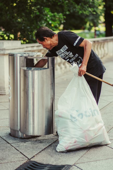 A man with dark hair, dressed in a black T-shirt with white printed text on the front, is seen disposing of rubbish into a stainless steel litter bin while holding a white plastic bag filled with waste. He is standing on a paved surface in an outdoor setting, with a background featuring lush, green trees and shrubs. The scene appears to be part of an organized waste disposal process, possibly related to an outdoor cleaning activity or public rubbish collection, with the man focusing on proper waste disposal in line with environmental standards. The stainless steel bin has a sleek, reflective finish, and is positioned along a stone balustrade or low wall, indicating a formal outdoor area or park environment. The lighting suggests daylight, contributing to a clear and natural atmosphere. This scene subtly reflects on the importance of maintaining public spaces through proper rubbish removal, aligned with private waste management or independent collection services like those offered by Rubbish Removal Finsbury Park.