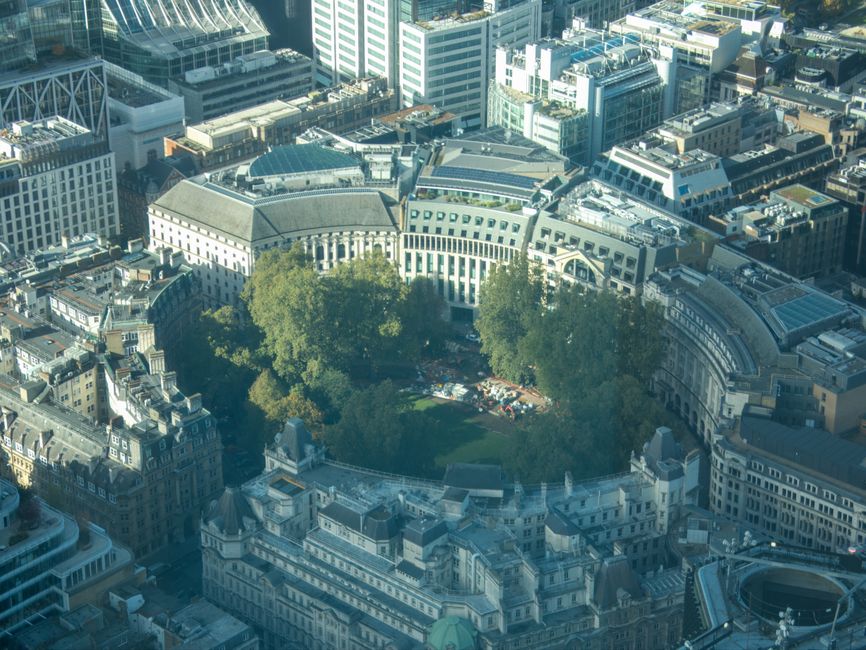 An aerial view of a circular park area surrounded by densely packed urban buildings, featuring numerous mature trees with green foliage occupying the central space. The park is enclosed by a variety of historic and modern architectural structures with ornate facades, flat roofs, and large windows. Some buildings have rooftop equipment such as HVAC units and skylights, indicating mixed-use urban development. Natural sunlight illuminates the scene, casting soft shadows and highlighting the textures of rooftops, facades, and tree canopies. The surrounding cityscape comprises high-rise office buildings, retail outlets, and residential blocks with varying heights and architectural styles. The overall environment appears busy yet well-maintained, showing a typical city center with limited visible activity at ground level. The image exemplifies an urban setting where private waste collection or on-site disposal could be relevant, especially within the context of independent rubbish removal services such as those provided by Rubbish Removal Finsbury Park, operating in the local area.