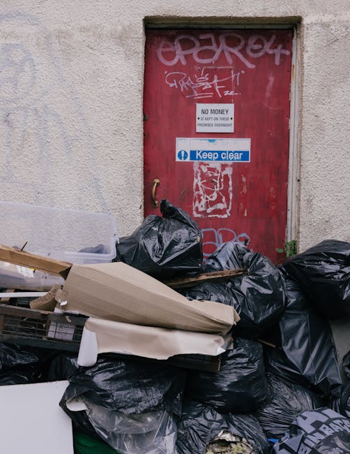 A collection of black plastic rubbish bags and discarded cardboard, some flattened, are piled in front of a red metal door on an exterior wall of a building. The door features various graffiti tags in white and pink, along with two signs: one indicating 'No Money - Please Do Not Leave Rubbish Overnight' and another blue sign stating 'Keep Clear.' The surrounding wall is textured and off-white, with signs of weathering and minor dirt. The rubbish appears to be accumulated in an unmanaged manner, suggesting a need for proper waste disposal, which an independent rubbish removal service from Rubbish Removal Finsbury Park could provide. The scene is illuminated by natural daylight, highlighting the contrast between the black bags, the red door, and the wall, with some greenery visible near the bottom right corner. This setting exemplifies typical urban waste accumulation that can be addressed through dedicated rubbish clearance services, ensuring a cleaner and more organized environment.