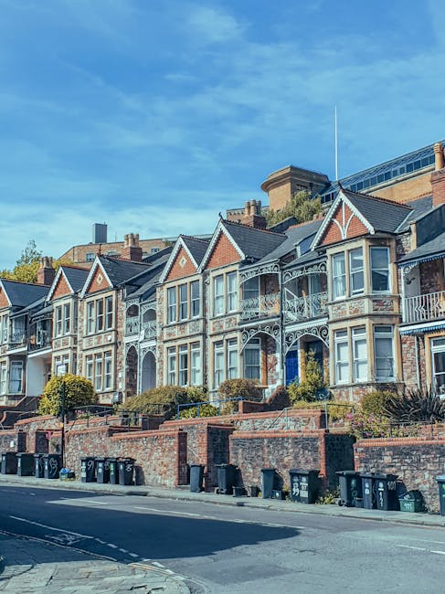 A row of historic Victorian-style terraced houses situated on a sloped street, featuring ornate white ironwork and decorative brick facades. The buildings have bay windows with white frames, gabled roofs with slate tiles, and small front gardens enclosed by low brick walls with metal railings. In front of the houses, there are black waste bins lined up along the pavement, indicating private rubbish collection or on-site clearance, which may be related to independent waste disposal services such as those provided by Rubbish Removal Finsbury Park. The sky above is partly cloudy with patches of blue, and the environment appears calm and residential. The street's curb and road surface are visible, and some greenery, including small shrubs, is present in the front gardens, contributing to the overall neighborhood aesthetic consistent with urban waste management considerations.