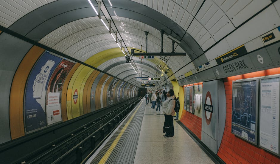 Inside the Green Park London Underground station, an arched underground platform with a curved tiled ceiling illuminated by linear lighting strips. The platform features a yellow tactile paving strip along the edge for safety, with a black metal railing separating the track area. On the right side, there are multiple large digital information screens and poster advertisements attached to the wall, which is covered in orange and grey tiles. Several commuters, some standing and others walking, are visible on the platform, dressed in casual attire and carrying backpacks or bags. Above, signs indicate directions and station information, including the Green Park station name displayed on a white sign with black text and the London Underground roundel logo. The environment appears clean, well-lit, and designed for efficient passenger movement, consistent with an urban underground metro setting where private and alternative travel options are often utilized for quick and convenient access to central London locations. This scene highlights how transport infrastructure supports daily commuting, aligning with services related to managing station waste and ensuring cleanliness in public transit environments, similar to the offerings of Rubbish Removal Finsbury Park for local waste handling and collection needs.
