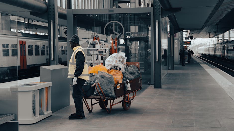 A man wearing a grey beanie, black jacket, and white gloves stands on a train station platform beside a large metal trolley filled with various types of rubbish, including plastic bags and orange-wrapped items. The trolley is positioned near a waste disposal station with a grey container to the man's left. Behind him, there are two train carriages, one white with red sliding doors and the other visible further along the platform. The platform has a tiled surface with a shelter featuring glass panels and metal supports, and there are a few other passengers in the background near the station entrance and train tracks. The scene appears to be during daytime with natural light illuminating the area, which is part of an urban train station environment. This image exemplifies a private or independent rubbish collection process at a transport hub, aligned with waste removal and disposal services provided by companies like Rubbish Removal Finsbury Park, ensuring the safe handling of discarded items while maintaining the cleanliness of public spaces.