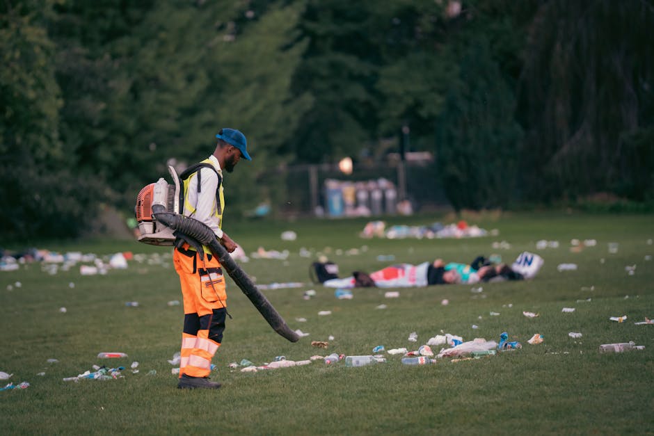 A male waste management worker wearing a blue cap, high-visibility jacket, and bright orange trousers with reflective stripes is using a leaf blower or vacuum to clear away litter from a grassy park area. The worker is standing in the foreground, holding a long, flexible hose attached to a backpack-style waste collection device. The ground around him is scattered with plastic bottles, paper cups, and other miscellaneous rubbish, indicating a recent incident of littering or a cleanup operation. In the background, there are two blurred figures lying on the grass, possibly emergency responders or volunteers, with trees and a small rectangular structure, such as a waste disposal station, further behind. The scene appears to be illuminated by natural daylight, providing clear visibility of the litter and the equipment, reflecting activities associated with alternative waste handling and private rubbish removal services by companies like Rubbish Removal Finsbury Park.