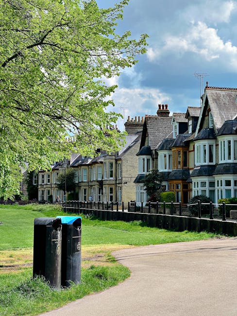 A residential street scene featuring a row of Victorian-style houses with varying rooflines, chimney stacks, and bay windows painted in neutral tones, with some painted in contrasting colours. In the foreground, there are two black public rubbish bins with blue lids, positioned on a grassy verge next to a concrete pavement. A large leafy tree with fresh green foliage extends its branches over the scene, providing partial shade. The background includes a partly cloudy sky with patches of blue visible through the clouds. The houses are separated from the pavement by low wooden fences, and the overall atmosphere suggests a quiet suburban area, typical for private waste management or independent rubbish collection methods supported by services like Rubbish Removal Finsbury Park.