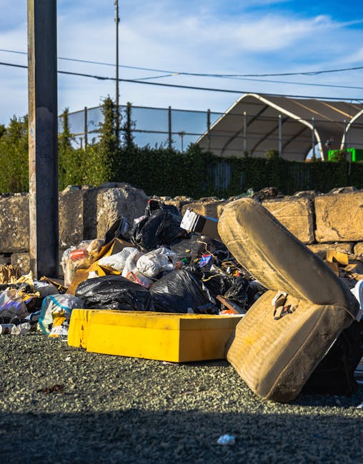 A male waste management worker wearing a blue cap, high-visibility jacket, and bright orange trousers with reflective stripes is using a leaf blower or vacuum to clear away litter from a grassy park area. The worker is standing in the foreground, holding a long, flexible hose attached to a backpack-style waste collection device. The ground around him is scattered with plastic bottles, paper cups, and other miscellaneous rubbish, indicating a recent incident of littering or a cleanup operation. In the background, there are two blurred figures lying on the grass, possibly emergency responders or volunteers, with trees and a small rectangular structure, such as a waste disposal station, further behind. The scene appears to be illuminated by natural daylight, providing clear visibility of the litter and the equipment, reflecting activities associated with alternative waste handling and private rubbish removal services by companies like Rubbish Removal Finsbury Park.